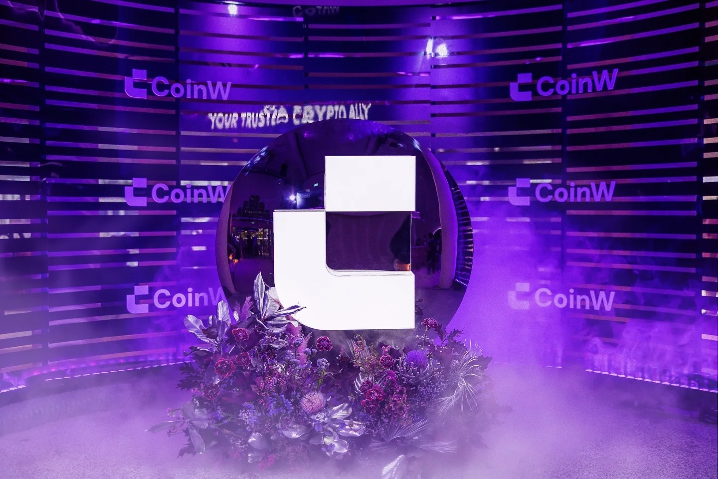 The CoinW logo set in flowers and low fog, in front of a wall of CoinW panels